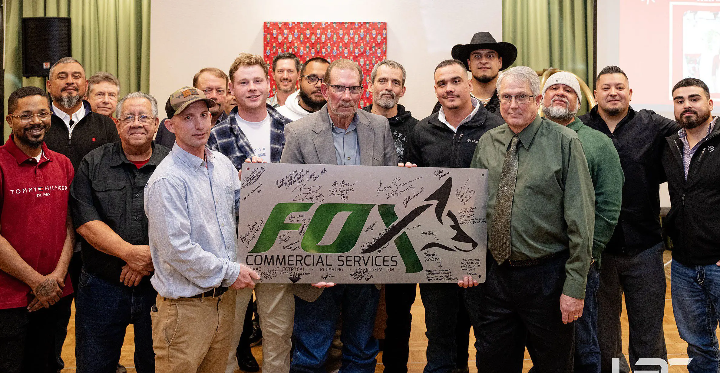 About LPC_History Section_Formarly Fox Commerc 2 A group of men pose indoors with a large Fox Commercial Services sign covered in signatures.