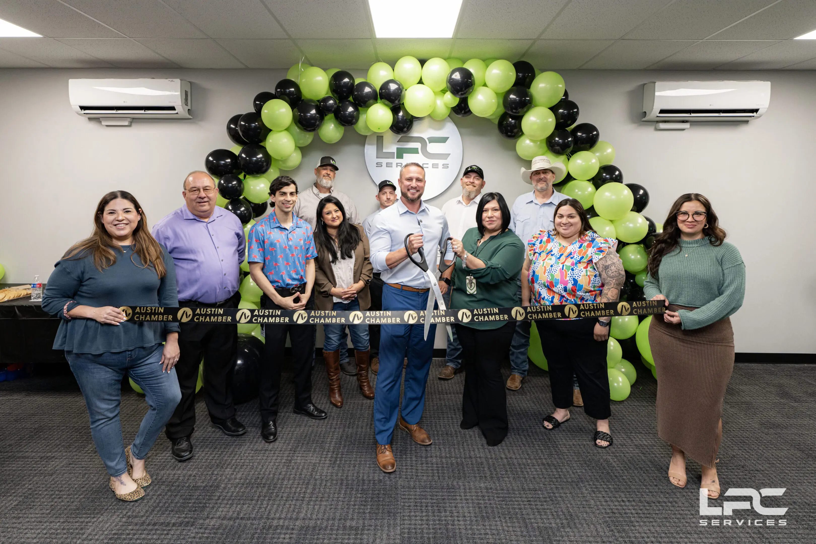 About LPC_Main Image_Ribbon Cutting A group of people pose indoors for a ribbon-cutting ceremony in front of a black and green balloon arch and a wall with an "LPC Services" logo.