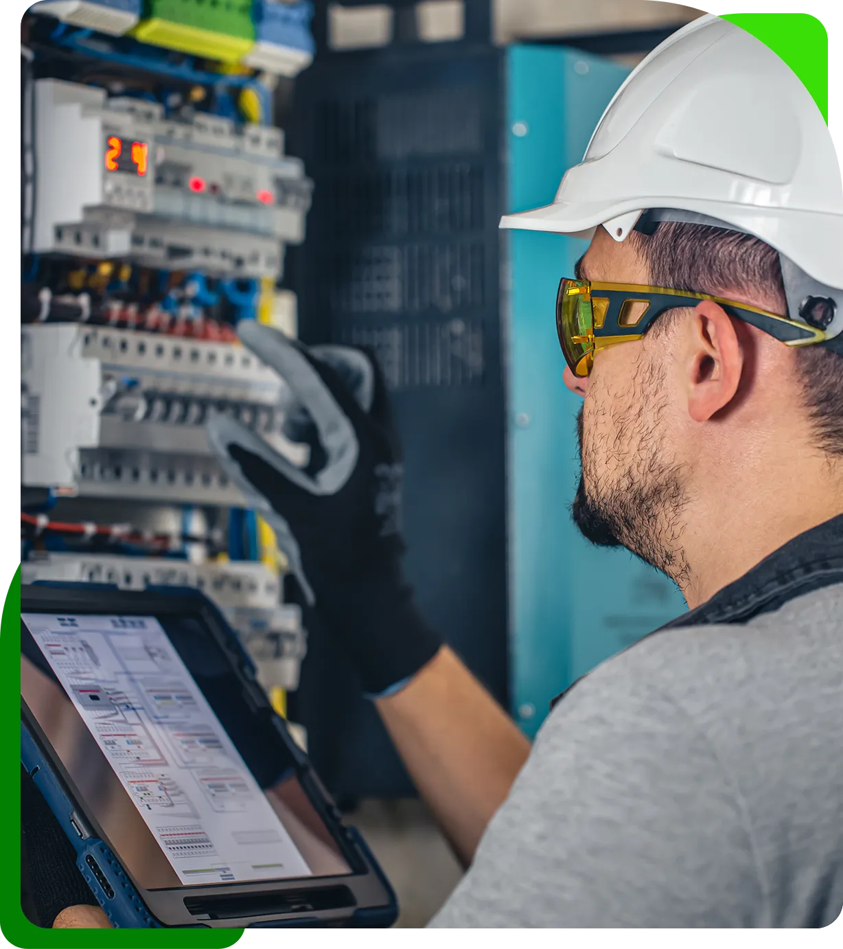 A worker wearing safety gear, including a helmet and goggles, uses a tablet to check electrical equipment with multiple switches and wires visible.