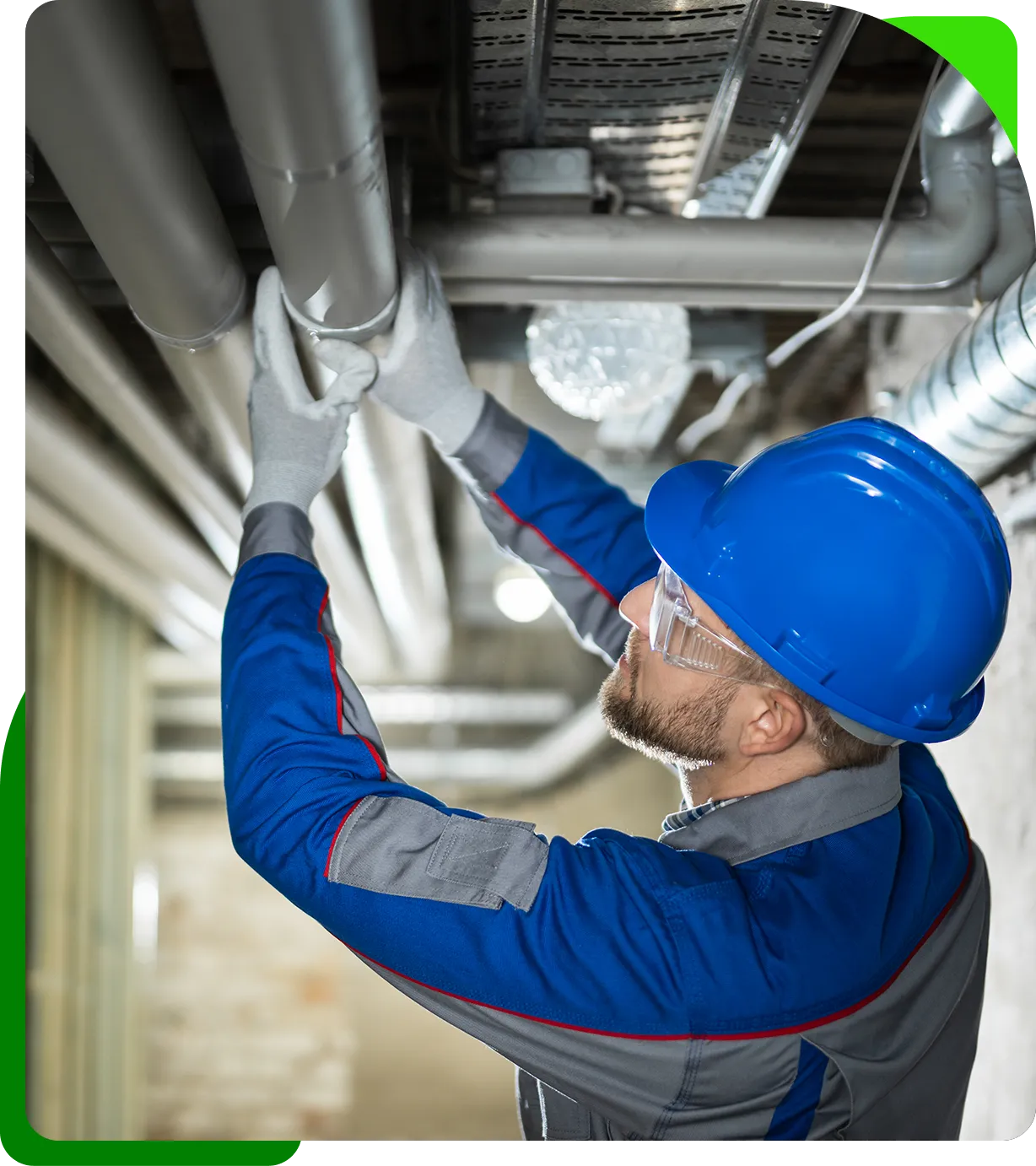 A worker wearing a blue hard hat and safety glasses inspects metal pipes on a ceiling, adjusting them with gloved hands.