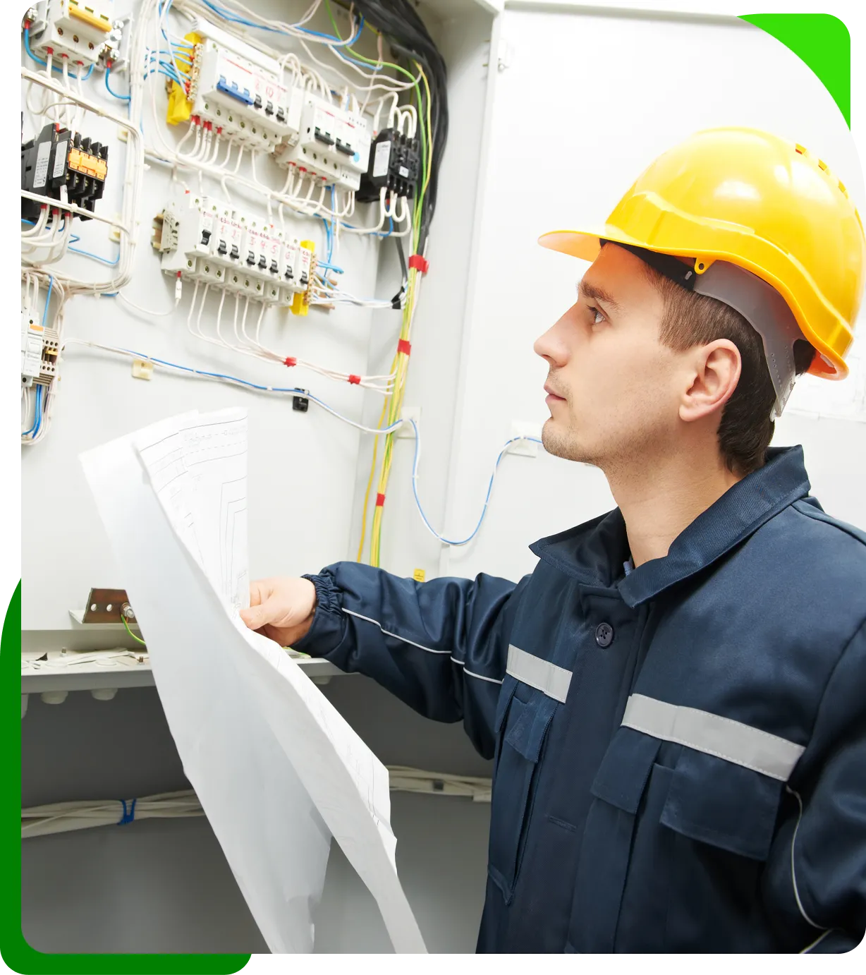 A person in a yellow hard hat examines electrical diagrams while standing in front of an open electrical panel.