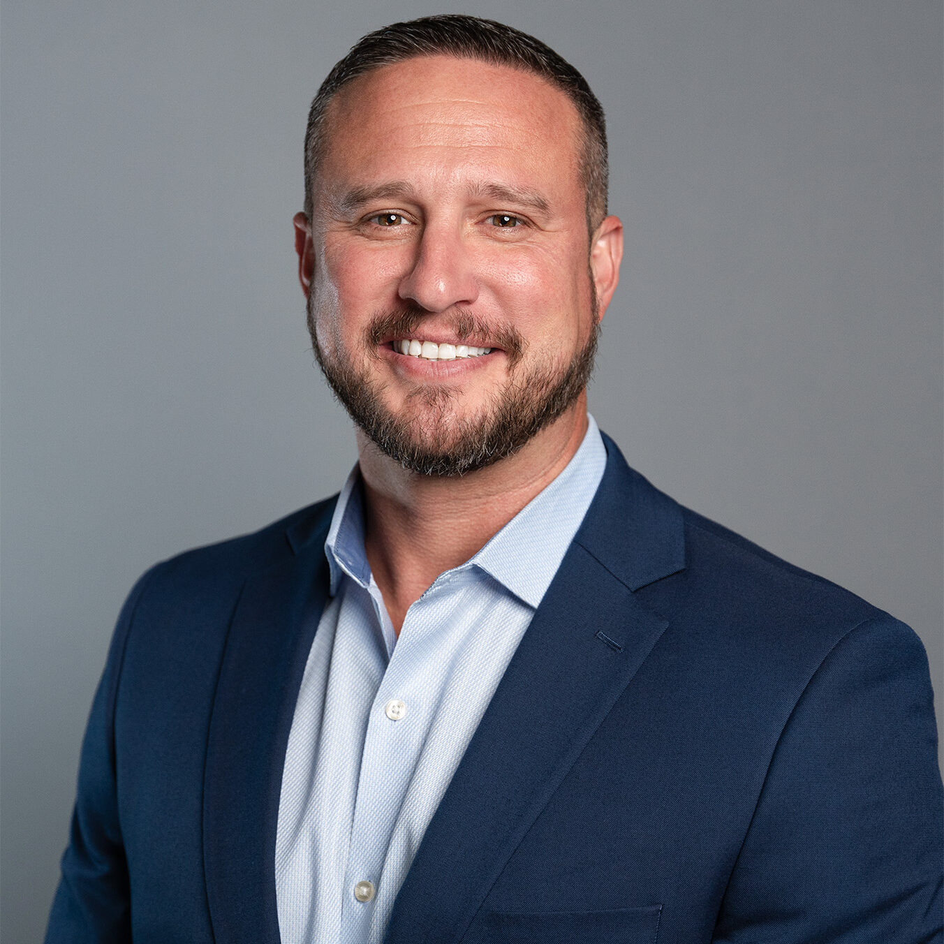 67f03ec914d94_About LPC_Meet The Team_Ben Milam A man in a blue suit and light blue shirt smiles at the camera against a plain gray background.