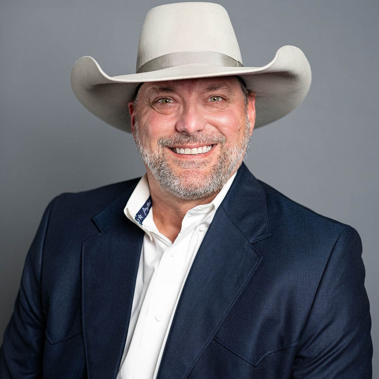 Brent Stroud A man wearing a light gray cowboy hat, navy blue blazer, and white shirt smiles in front of a plain gray background.