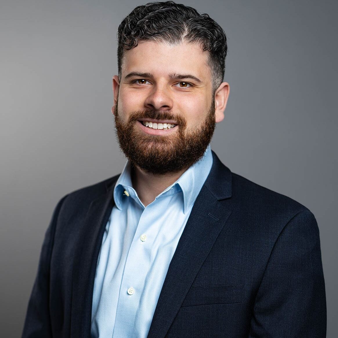 Chris Silva A man with short, curly dark hair and a beard, wearing a navy suit jacket over a light blue dress shirt, poses against a plain gray background.