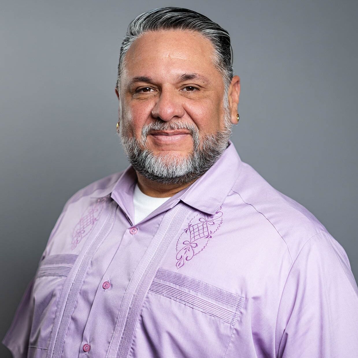 Daniel Villanueva A man with short gray hair and a beard smiles at the camera, wearing a light purple embroidered shirt, against a plain gray background.