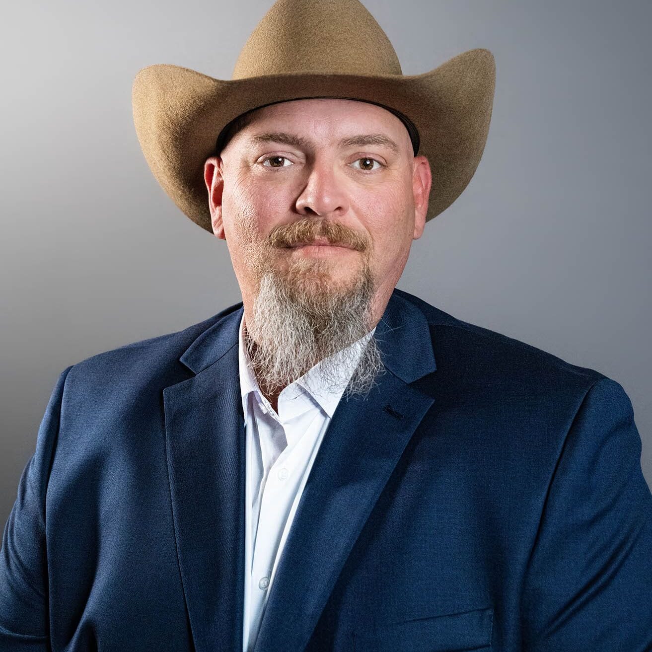 Dominick Dalessio A man with a light beard and mustache wearing a tan cowboy hat, blue suit jacket, and white shirt poses in front of a plain gray background.