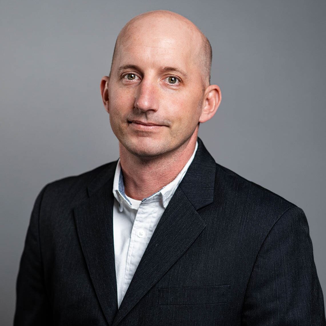 Jeff Brock A man in a dark suit and white shirt poses for a professional headshot against a plain gray background.