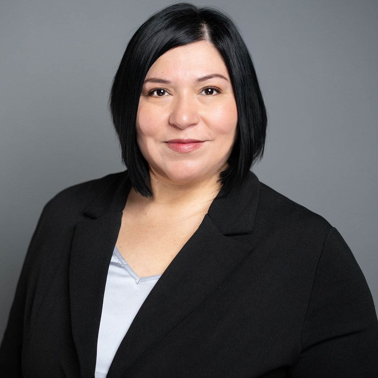 Lori Trujillo A woman with straight black hair in a bob cut, wearing a black blazer over a light gray top, poses against a plain gray background.