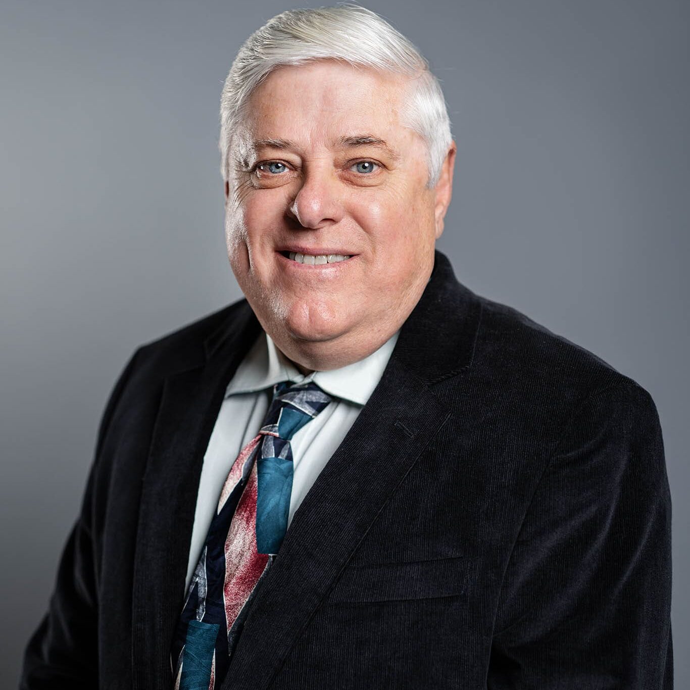 Mickey Foster Older man with short white hair wearing a dark suit jacket, light shirt, and patterned tie, standing in front of a plain gray background.