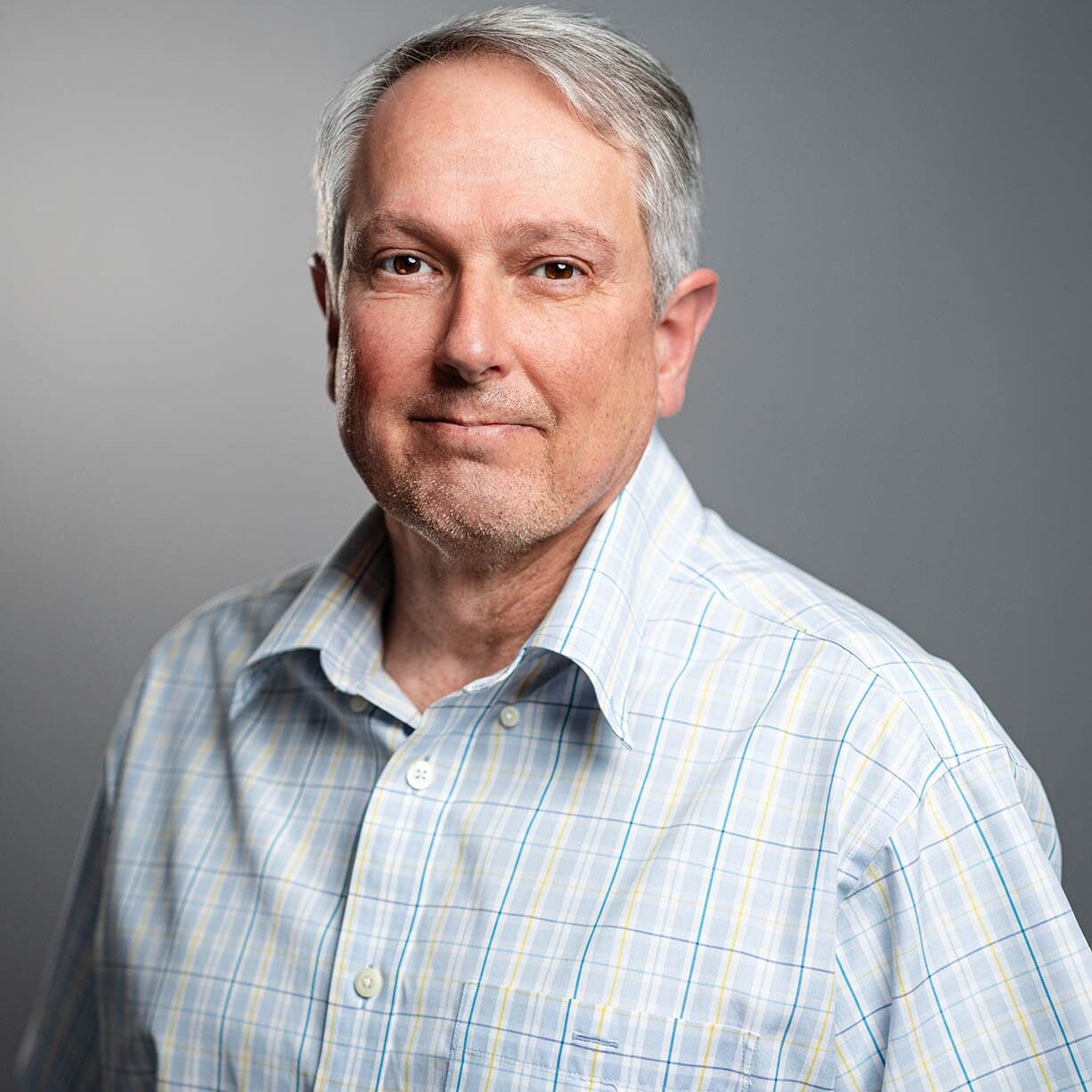 Stacey Warren A middle-aged man with short gray hair, wearing a light-colored, plaid button-up shirt, stands in front of a neutral gray background.